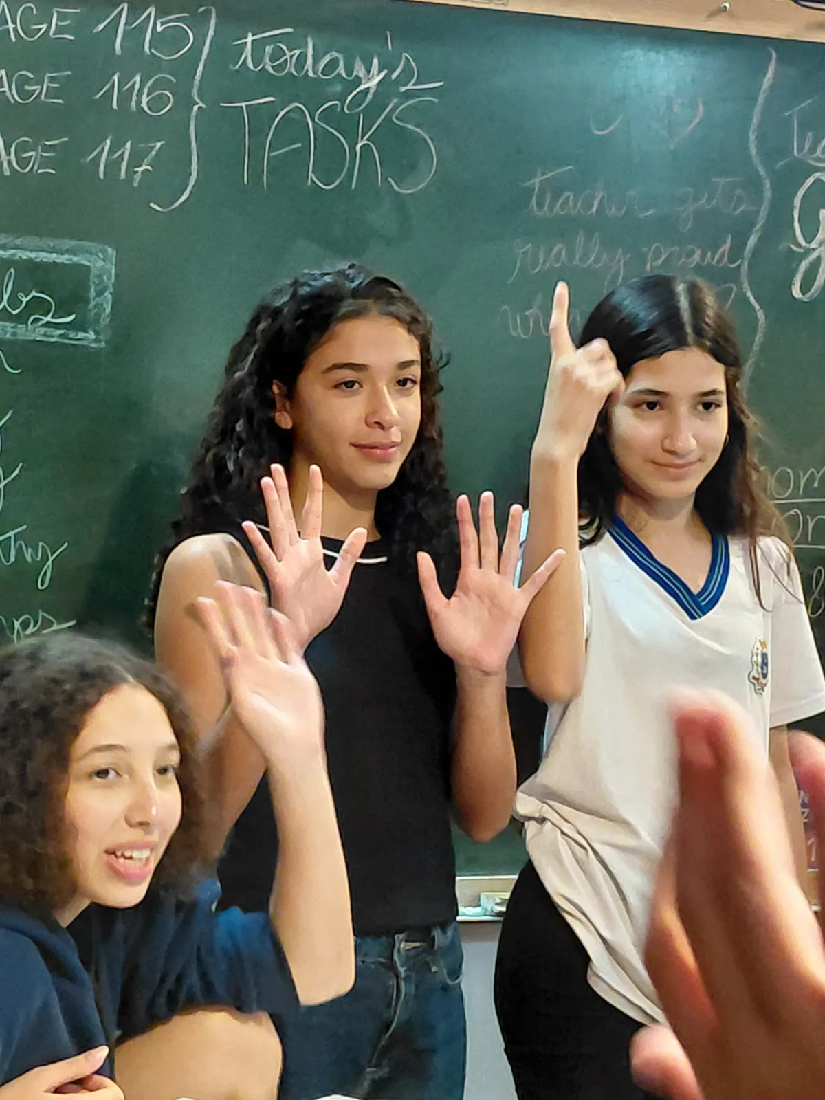 Três adolescentes sorrindo em frente ao quadro durante a aula de inglês